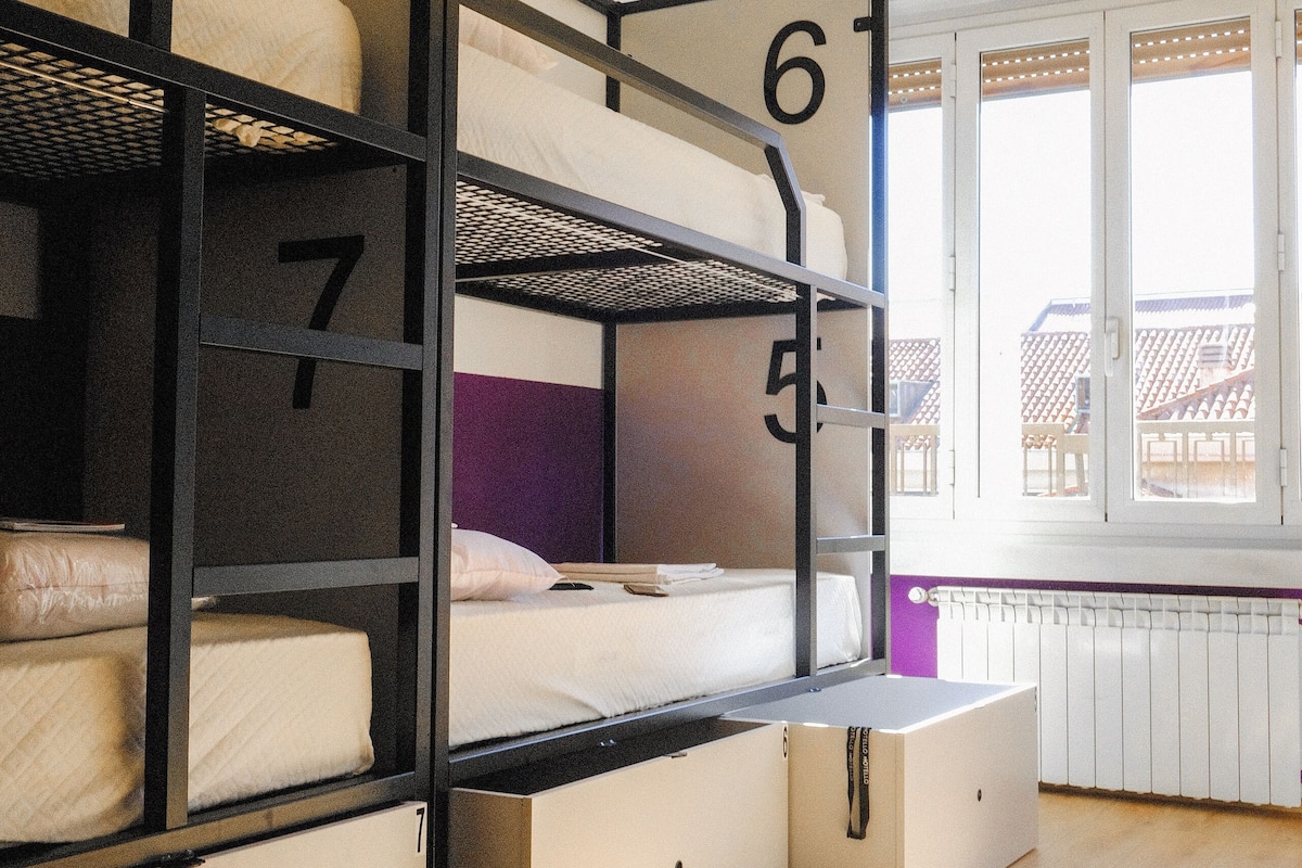 Une chambre avec des lits superposés, portant les numéros 6, 7 et 8, un plancher en bois et une fenêtre donnant sur des immeubles.