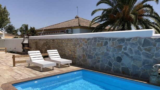 A swimming pool with two white lounge chairs, a stone wall, and a tiled deck. - Rocamar House (Barbate)