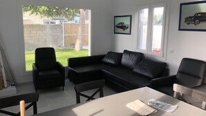 A modern living room with a black leather sofa, a dining table, and two chairs. There are framed pictures on the walls and a large window providing a view of the backyard. - Rocamar House (Barbate)