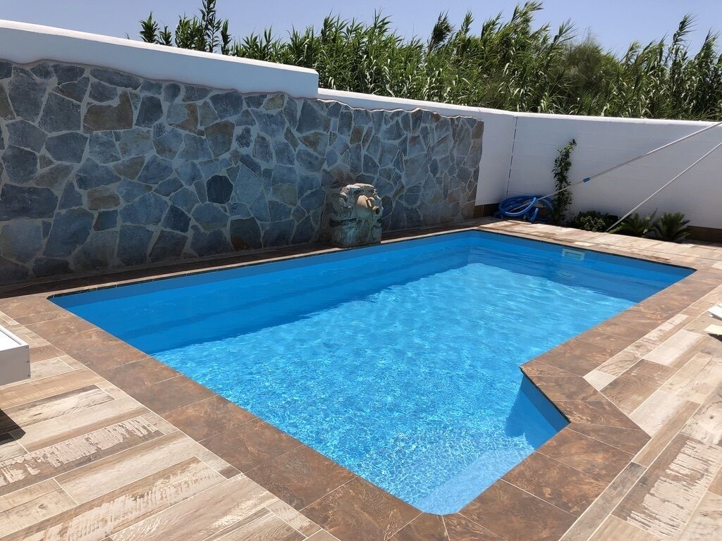 Una piscina rectangular con pared de piedra y solárium con baldosas.