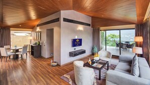 Una habitación de hotel moderna con techo de madera, una zona de comedor con mesa y sillas, un sofá y un ventanal con vistas al exterior.