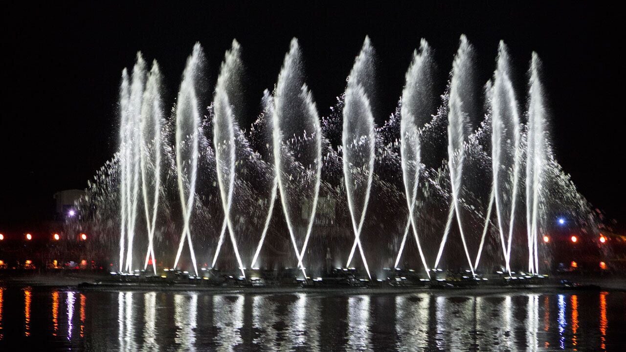 A nighttime view of a water fountain with multiple jets of water illuminated by lights, reflecting on the water surface.