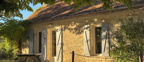 A rustic stone building with a tiled roof, wooden shutters, and a picnic table outside.