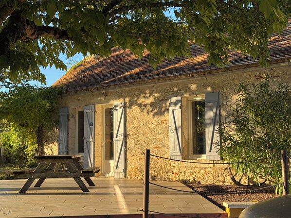 A rustic stone building with a tiled roof, wooden shutters, and a picnic table outside.