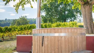 Een houten jacuzzi met een metalen schoorsteen, omringd door een betegeld terras, een houten bank en een rode muur.