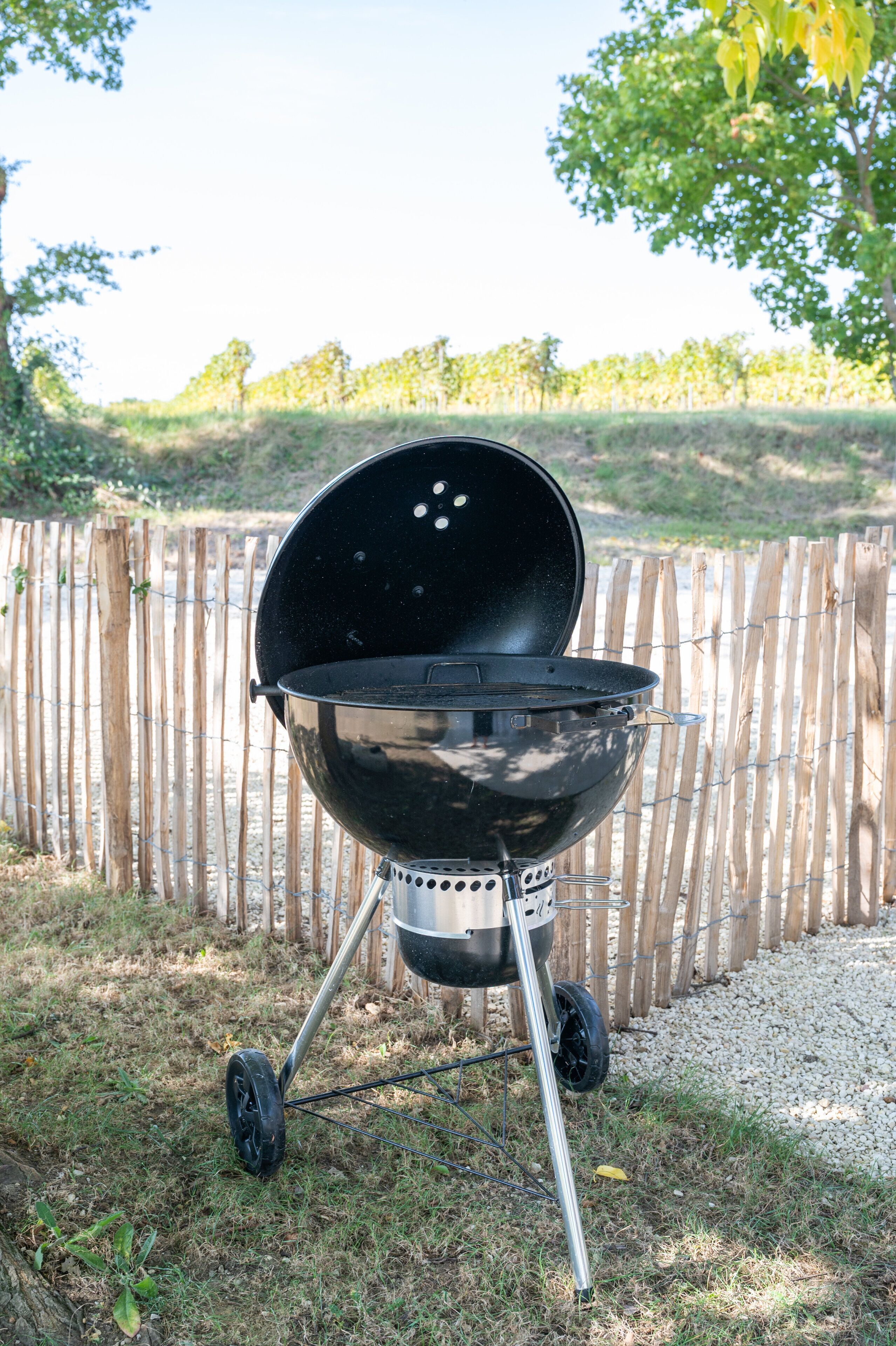 Un barbecue noir pour l’extérieur, doté d’un couvercle, de roues et d’une structure en métal, placé sur une pelouse, avec une clôture en bois en arrière-plan.