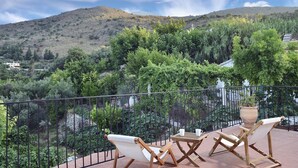 Eine Terrasse mit zwei Holzliegen und einem Tisch, mit Blick auf einen üppigen Garten und eine Hügelkuppe.
