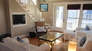 A living room with a staircase, white furniture, a glass coffee table, and a lit table lamp. - Sandcastle by the Sea - 3Bedroom, 2 Bath/Sleeps 6 - South Beach - Oceanside  (Bald Head Island)