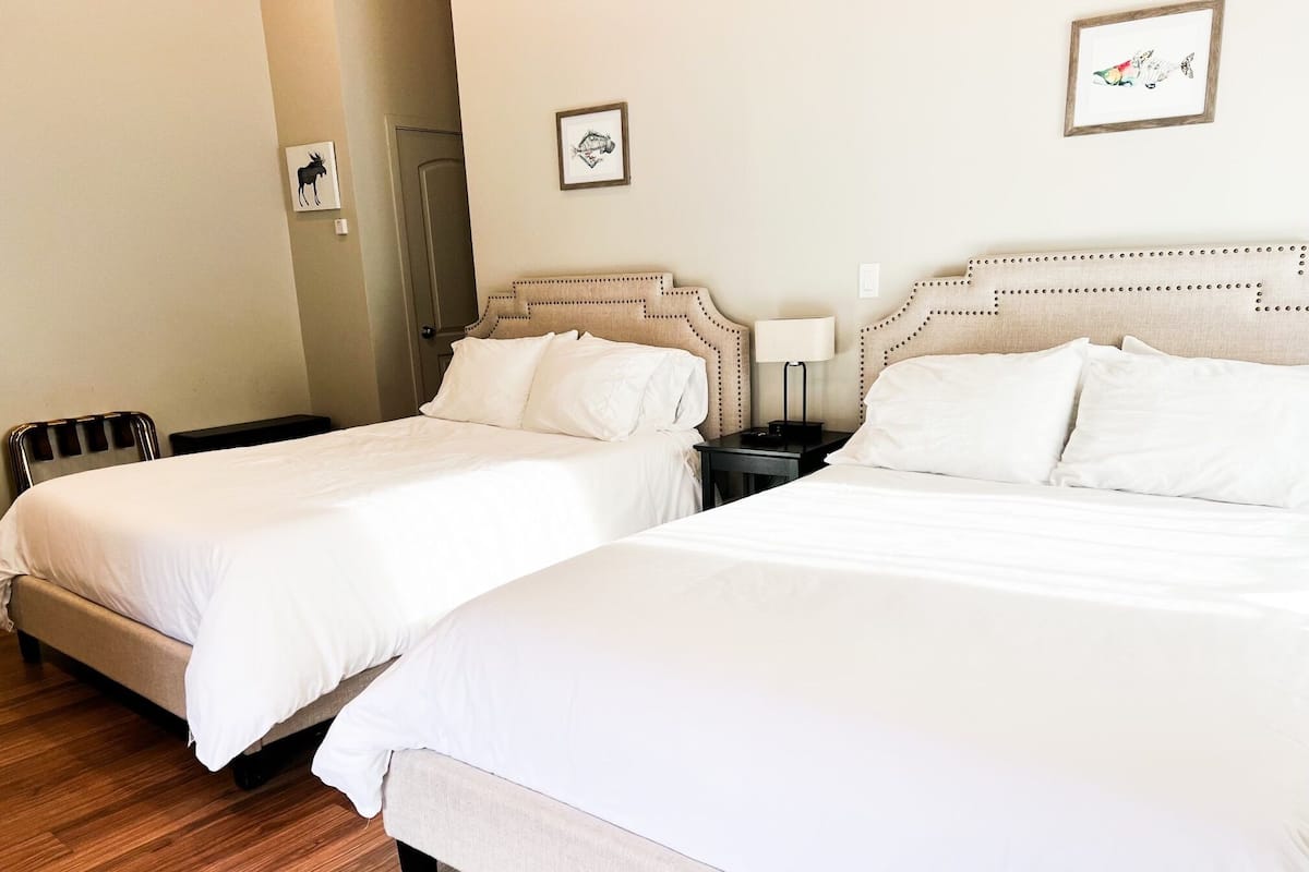 Two single beds with white linens, a wooden floor, and a beige wall with two framed pictures.
