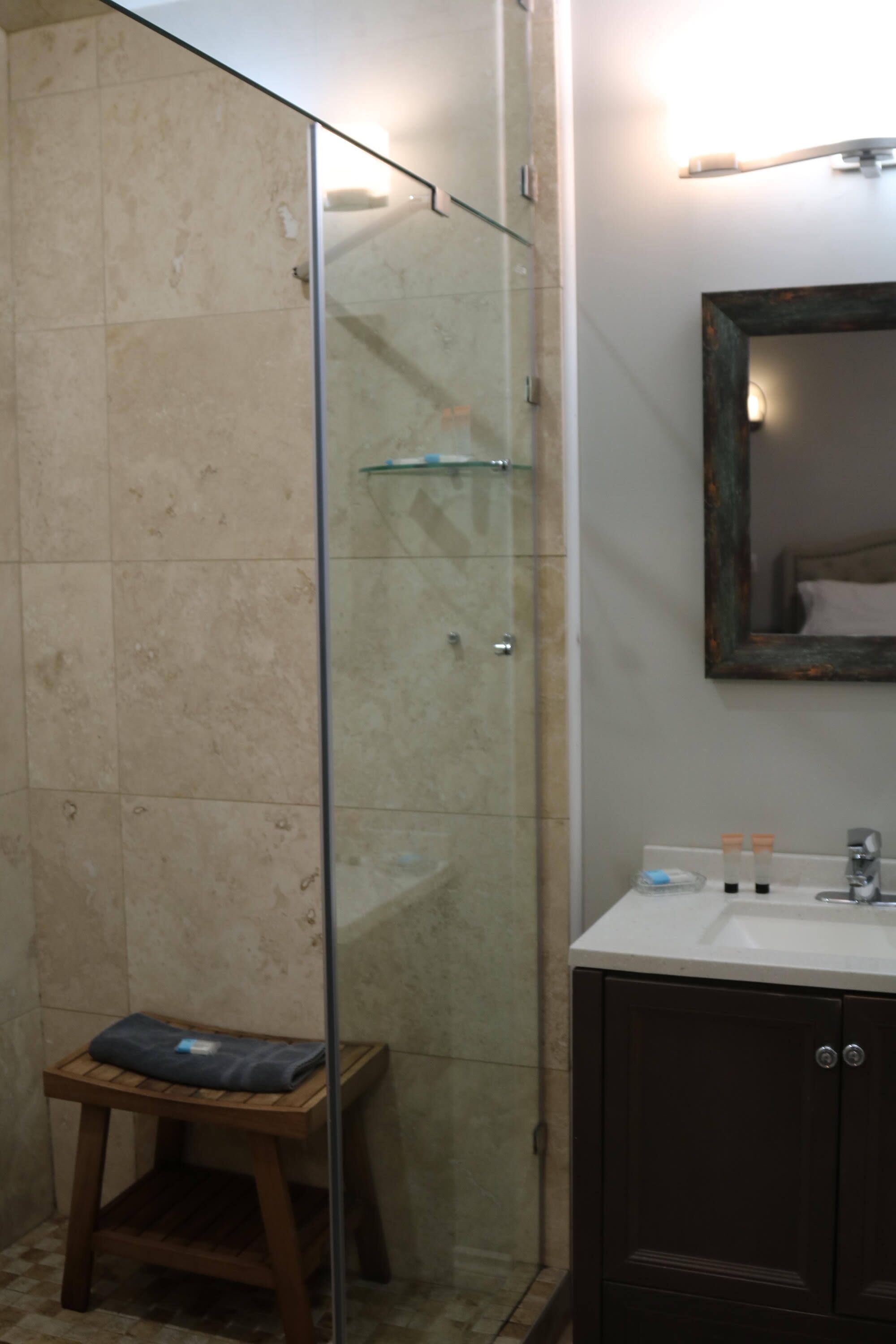 Une salle de bain avec une douche à parois de verre, un tabouret en bois, un lavabo avec des articles de toilette et un miroir doté d’un cadre décoratif.