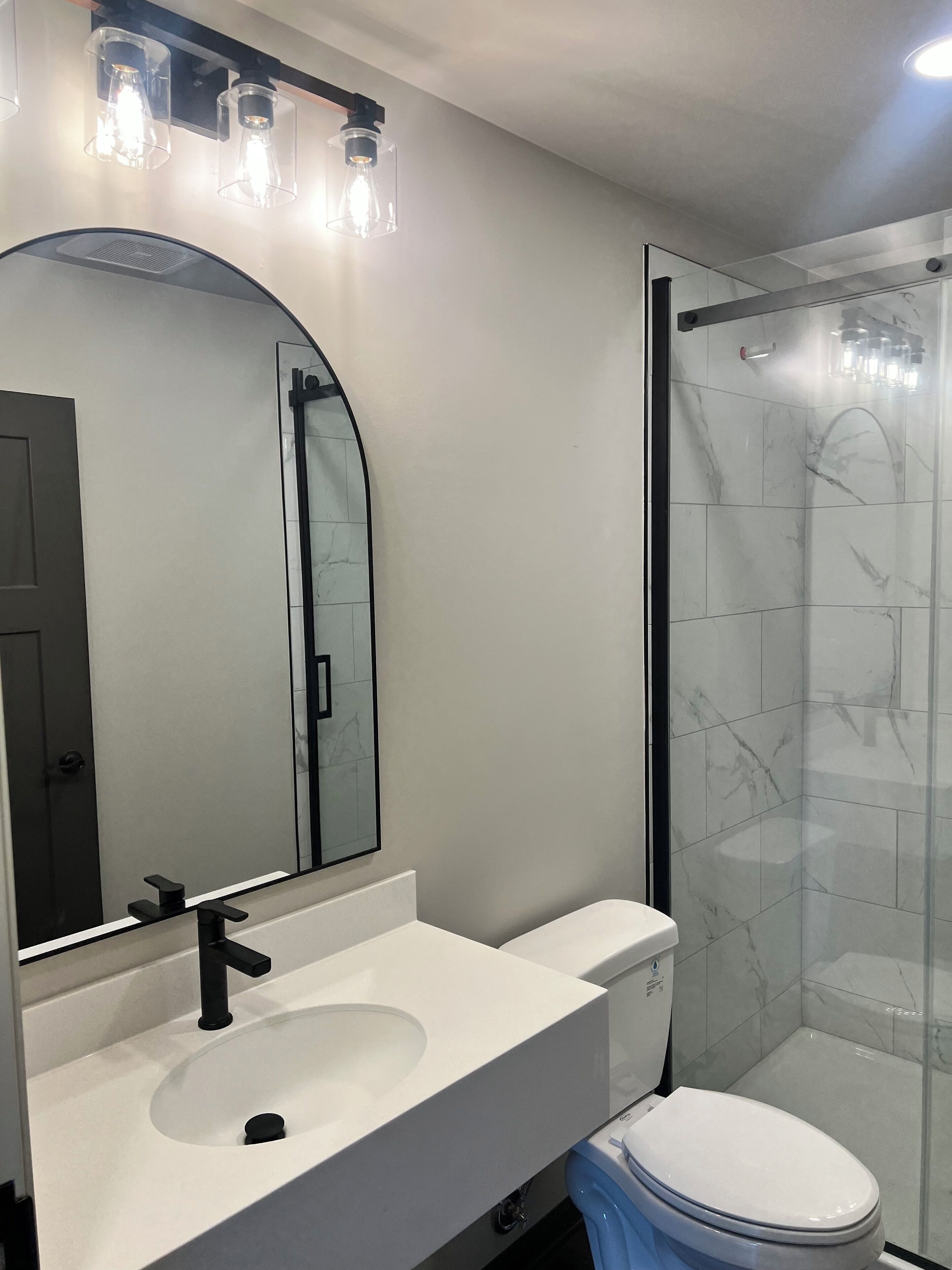 A modern bathroom with a white sink, black faucet, and a large mirror. There is a glass shower enclosure with a marble tile wall.