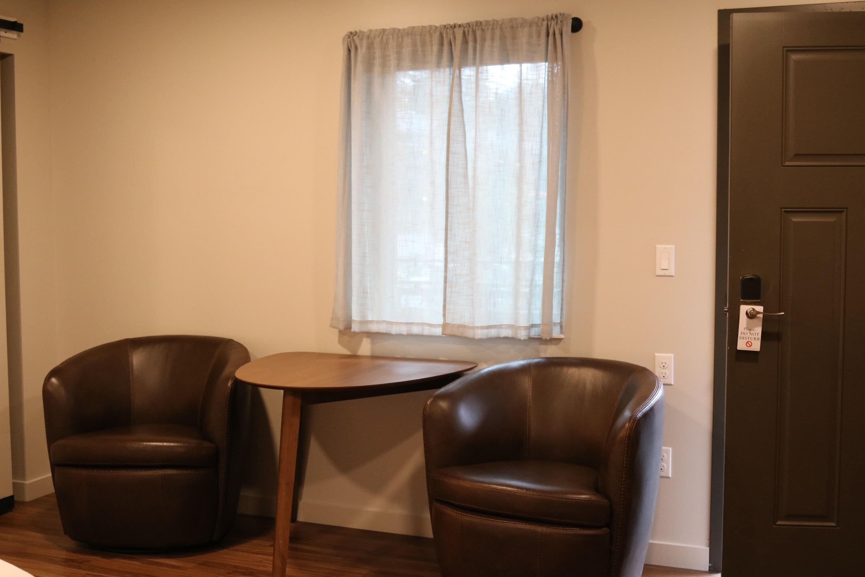 Une pièce comprenant deux fauteuils en cuir, une petite table ronde, une fenêtre ornée de rideaux légers et une porte dotée d’une serrure.