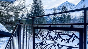 Une terrasse en bois avec une rambarde en métal, d’où l’on peut admirer un paysage montagneux enneigé.