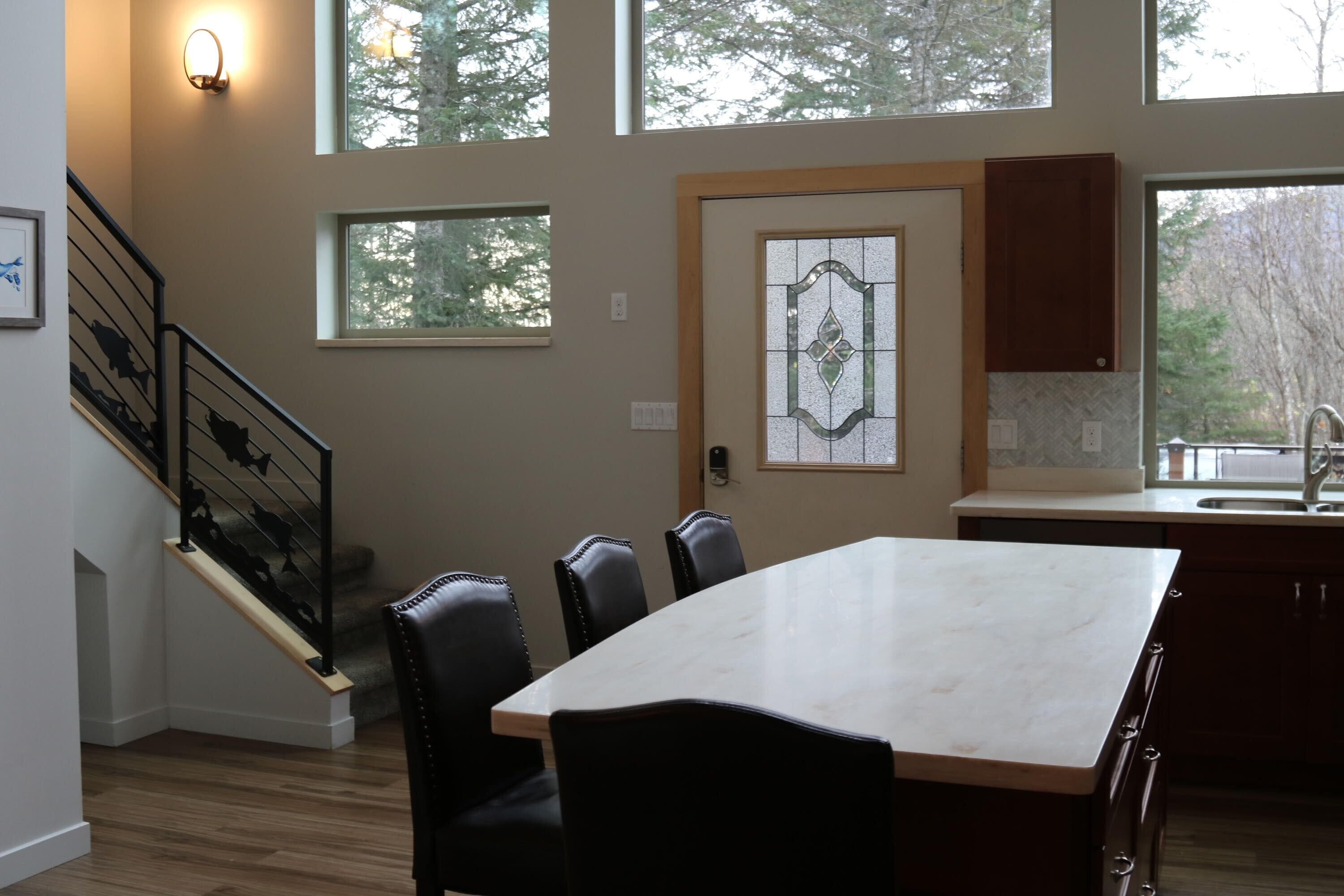 A modern kitchen with a large island, a dining area with chairs, a staircase, and a view of trees through the windows.