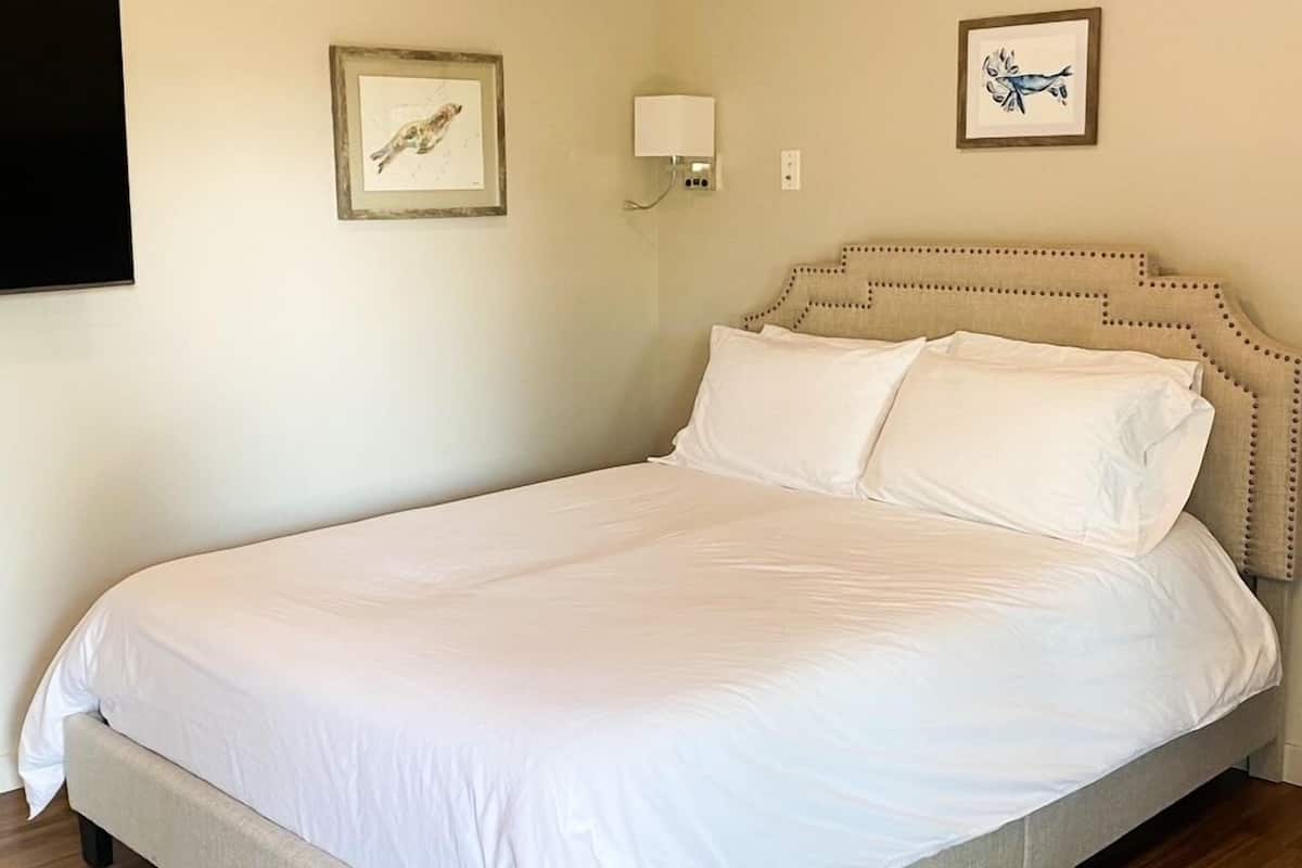 A neatly made bed in a room with a wooden floor, a ceiling light, and two framed pictures on the walls.