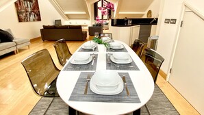 Un espace repas avec une longue table dressée pour quatre personnes, entourée de chaises modernes, dans un appartement de style loft doté de hauts plafonds et offrant une vue sur le ciel.