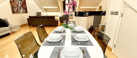 A dining area with a long table set for four, surrounded by modern chairs, in a loft-style apartment with high ceilings and a view of the sky.