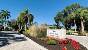 A sign for '500 Aldea Mar Village by the Sea' with a paved pathway and landscaped area with palm trees and red flowers. - Stand alone villa  (Venice)