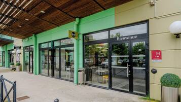 L’entrée d’un hôtel, avec une façade verdoyante, une enseigne indiquant « Bienvenue » et une entrée accessible aux personnes à mobilité réduite.