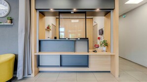 A reception area with a wooden desk, a clock on the wall, and a potted plant. - B&B HOTEL Cambrai (Proville)