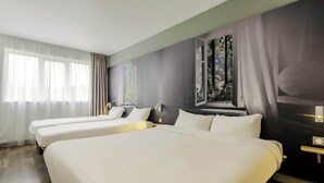 A hotel room with three single beds, a large window with a view of trees, and a wall-mounted nightstand with a lamp. - B&B HOTEL Cambrai (Proville)