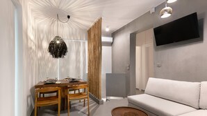A modern living room with a white sofa, a dining table with chairs, a hanging light fixture, and a mounted TV. - Elisa Seaside Luxury Suite Private hot Tub, Beach (Loutraki-Agioi Theodoroi)