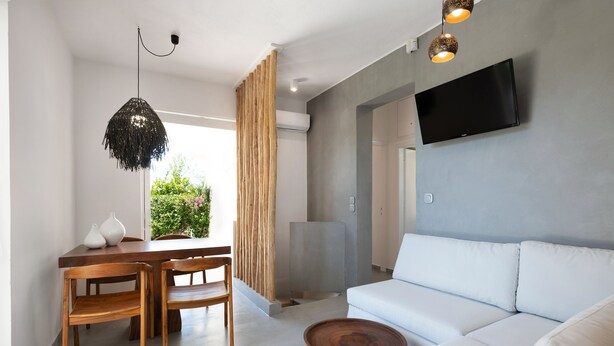 A modern living room with a white sofa, wooden dining table, and chairs. A flat-screen TV is mounted on the wall, and there is a large window with a view of greenery. - Elisa Seaside Luxury Suite Private hot Tub, Beach (Loutraki-Agioi Theodoroi)