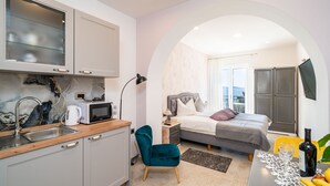 A compact hotel room with a kitchenette, a bed with a grey headboard, a small sitting area, and a view through an arched doorway. - Apartments Villa Palma (Zupa dubrovacka)