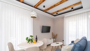 A modern living room with a blue sofa, a dining table with chairs, a TV mounted on the wall, and a large window with sheer curtains. - Apartments Villa Palma (Zupa dubrovacka)