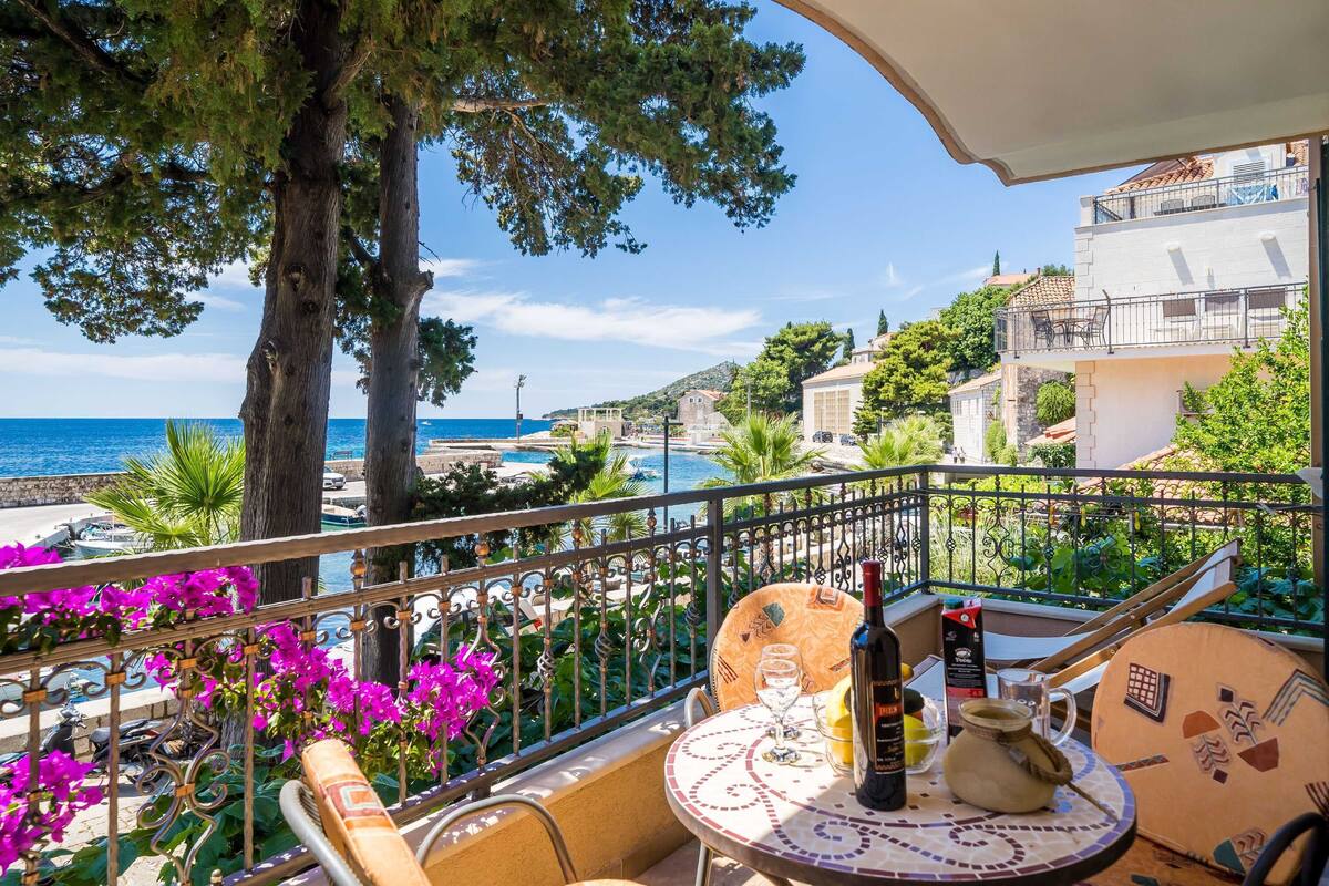A balcony with a table, chairs, and a bottle of wine. There are trees and a view of the sea and buildings in the background.