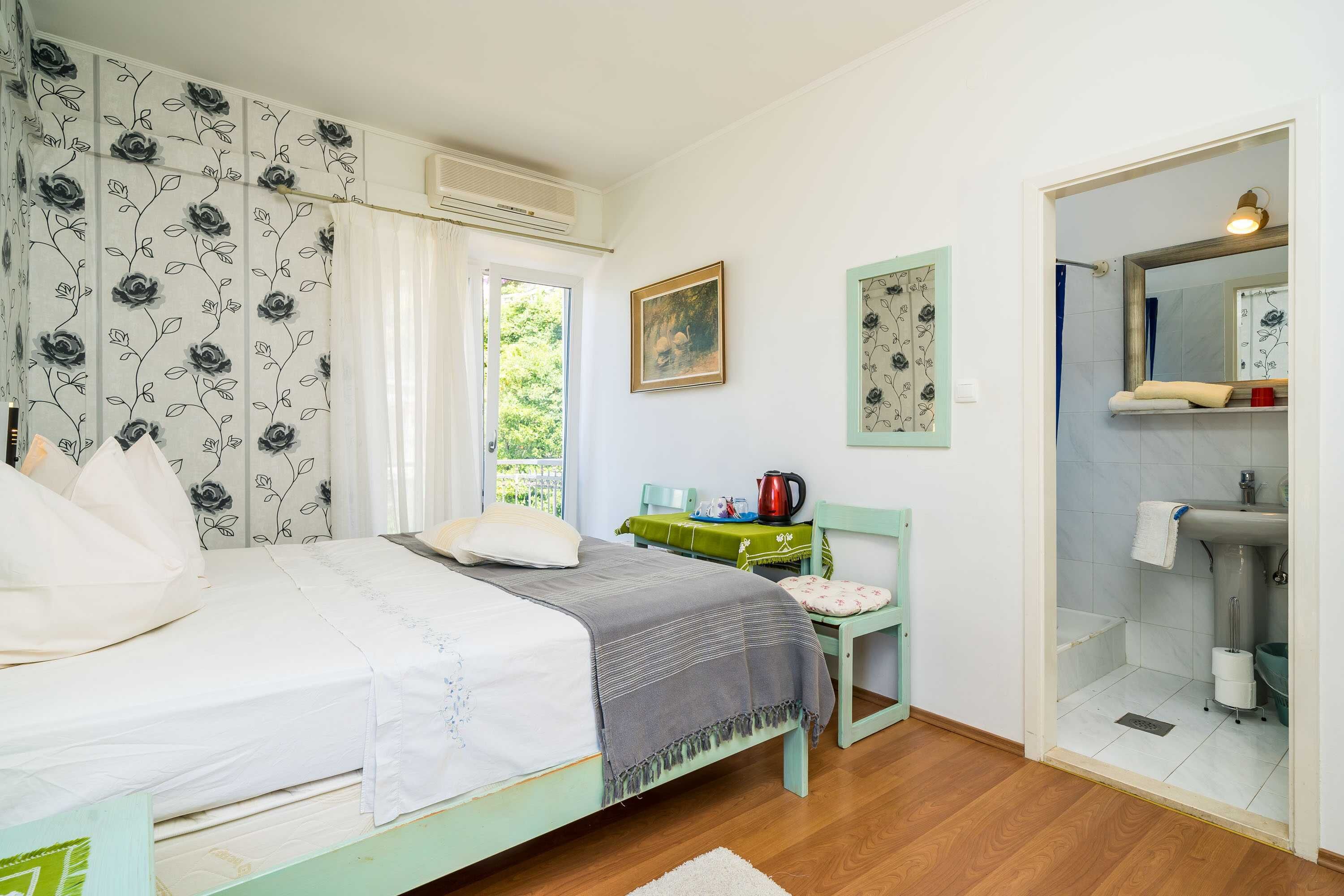 A bedroom with a bed, a chair, a table with a kettle, and a bathroom with a sink and mirror.