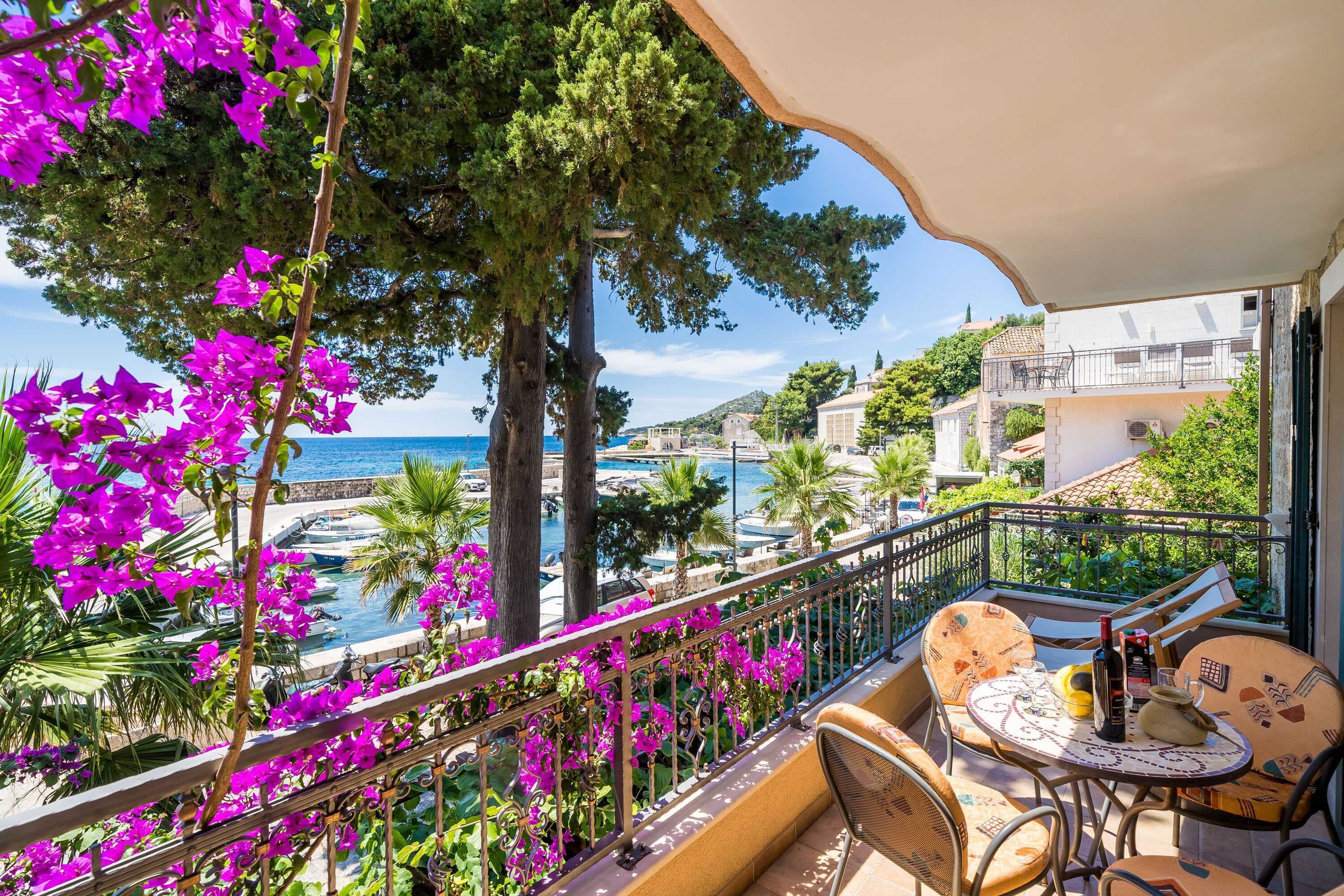 A balcony with a view of the sea, surrounded by vibrant bougainvillea flowers and a palm tree.