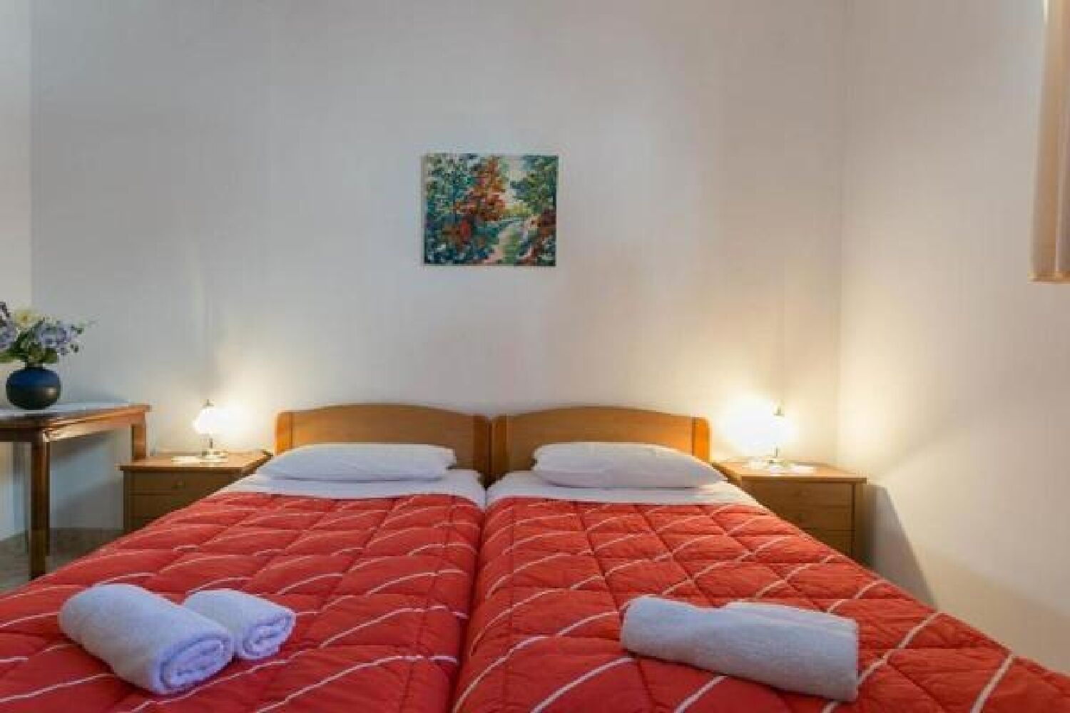 Een tweepersoonsbed met een rode gewatteerde sprei en twee opgerolde handdoeken. Twee nachtkastjes met lampen, een klein schilderij aan de muur, en een vaas met bloemen aan de linkerzijde.