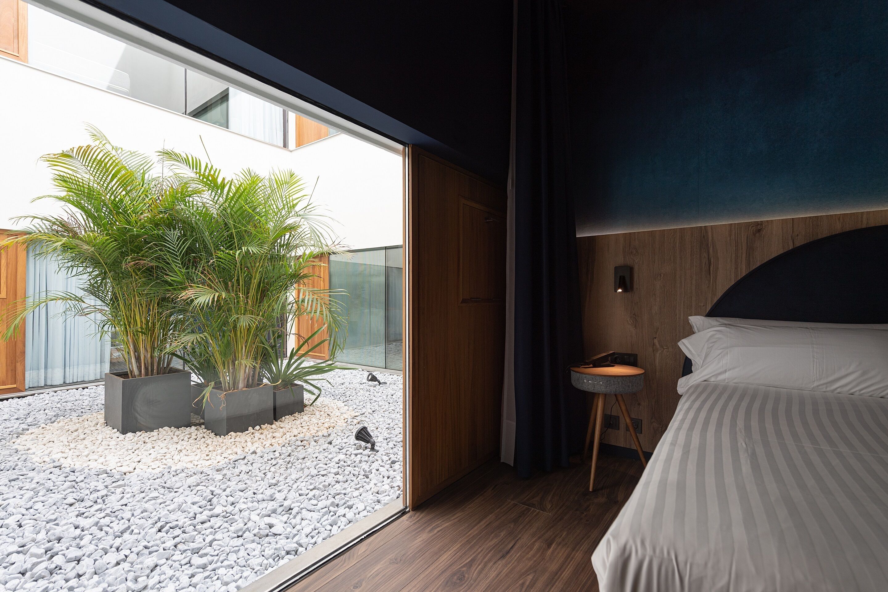 Un dormitorio moderno con un ventanal grande, una cama, una mesita de noche pequeña y una planta en maceta.