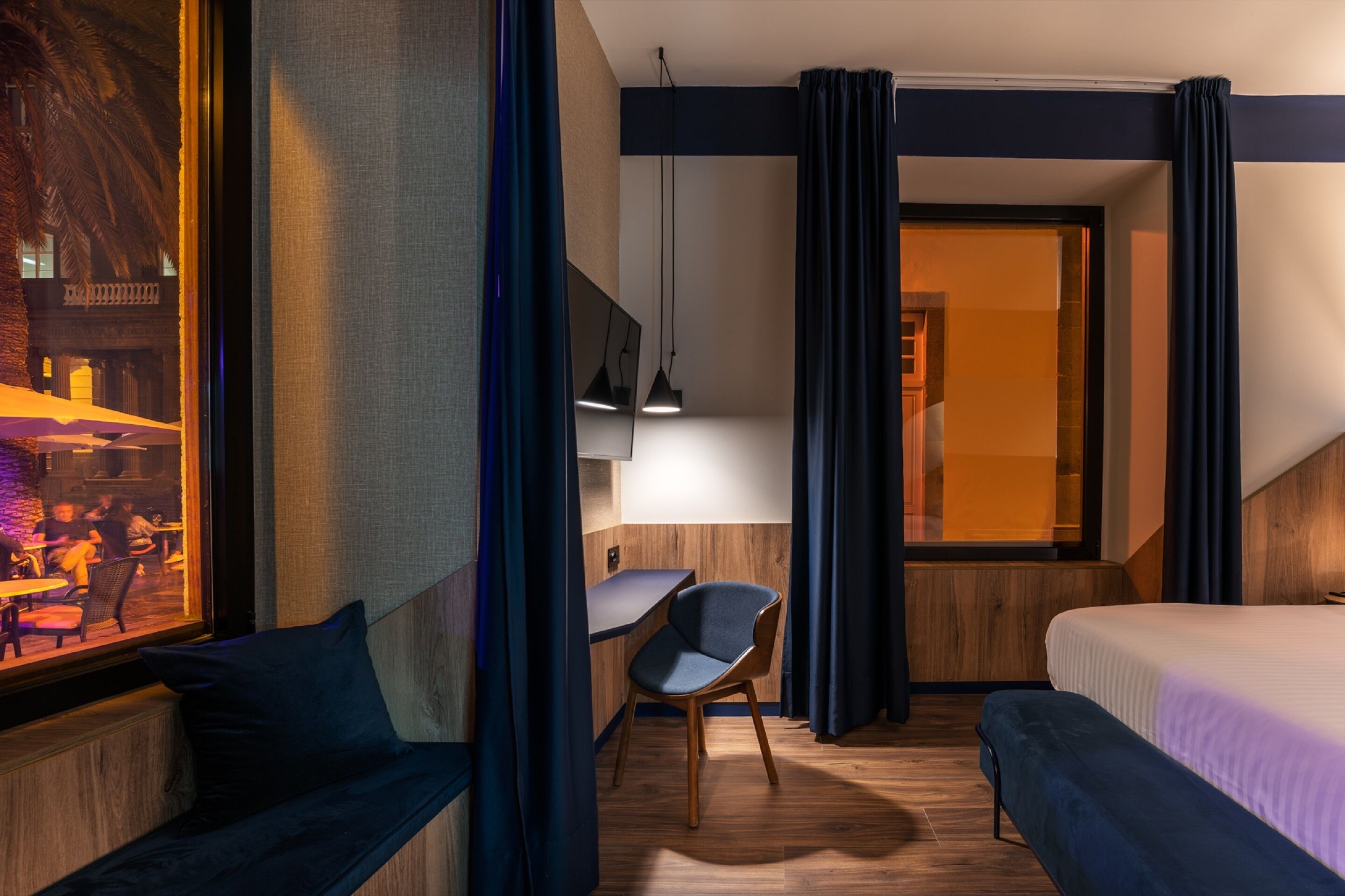 Ein modernes Hotelzimmer mit Bett, Schreibtisch und Stuhl. Es gibt ein Fenster mit Vorhängen, eine Hängelampe und Blick auf ein Restaurant vor dem Haus.