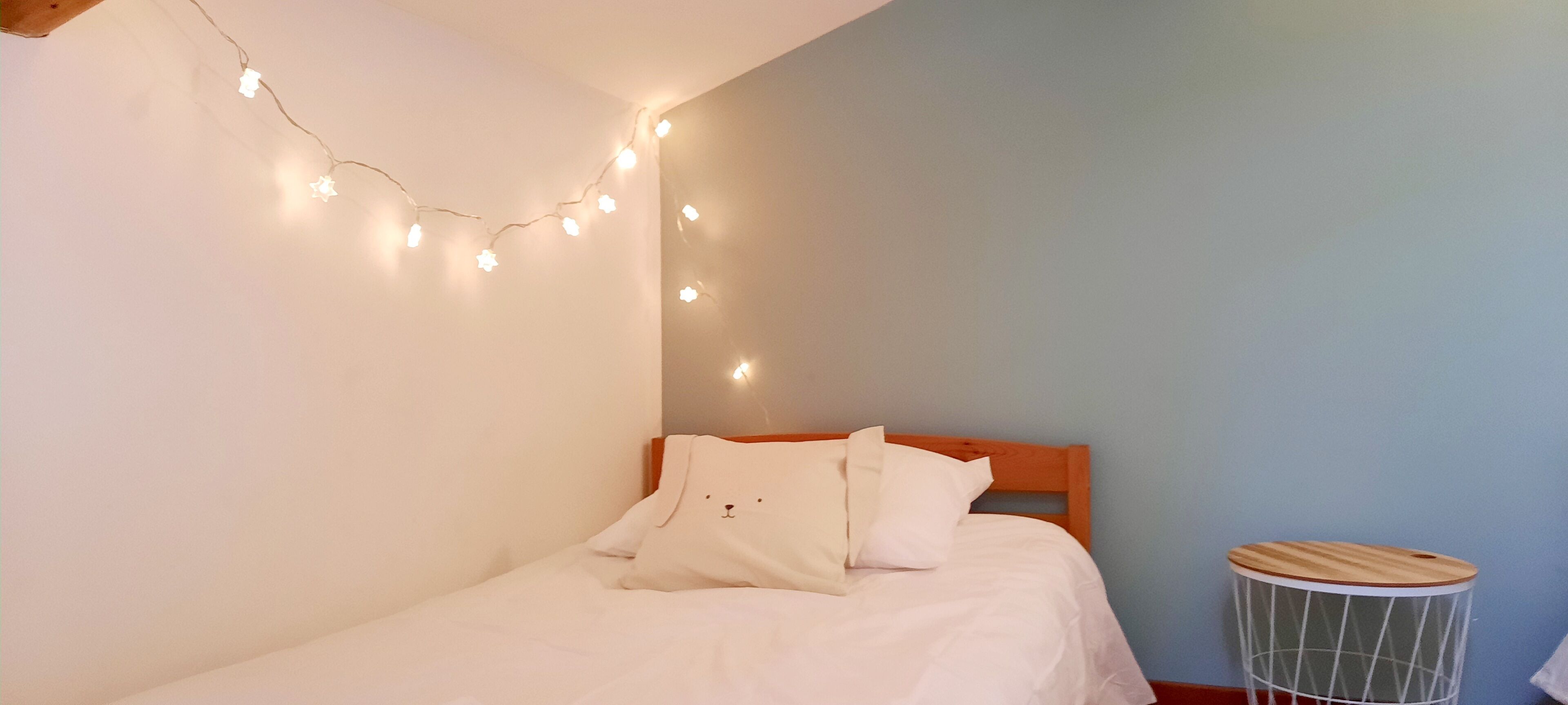 A bedroom with a bed, a wooden headboard, a bedside table, and string lights on the wall.