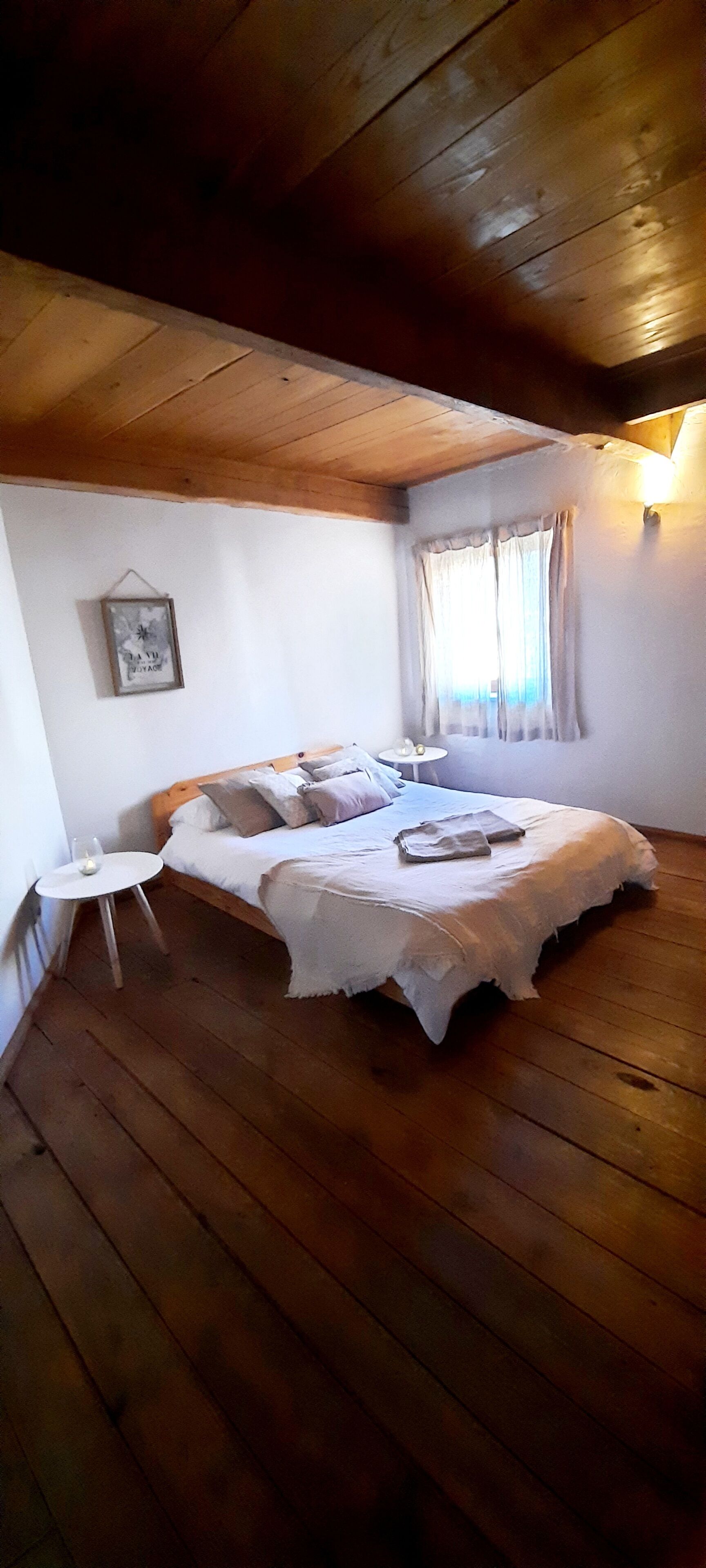 A bedroom with a wooden ceiling, a bed with a white bedspread, a small round table with a glass and candle, and a framed picture on the wall.