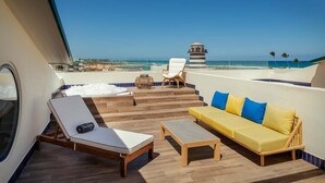 A sunlit outdoor deck with a yellow sofa, white lounge chairs, and a small table. A clear view of the ocean and a distant building is visible. - Ocean El Faro - Privilege RoofTop Junior Suite Pool view (Punta Cana)