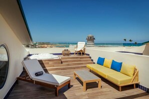 A sunlit outdoor deck with a yellow sofa, white lounge chairs, and a small table. A clear view of the ocean and a distant building is visible. - Ocean El Faro - Privilege RoofTop Junior Suite Pool view (Punta Cana)