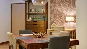Un coin repas comprenant une table en bois, des chaises, un élément décoratif central, un mur recouvert d’un papier peint à motifs et une lampe.