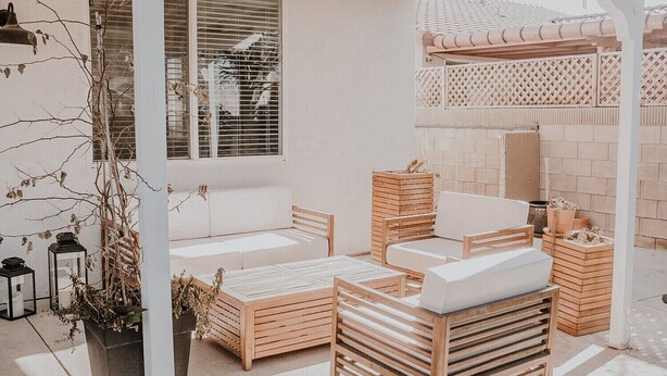 Un espace patio aménagé avec un canapé blanc, des tables et des chaises en bois. On y trouve également des plantes en pot et une petite structure en bois.