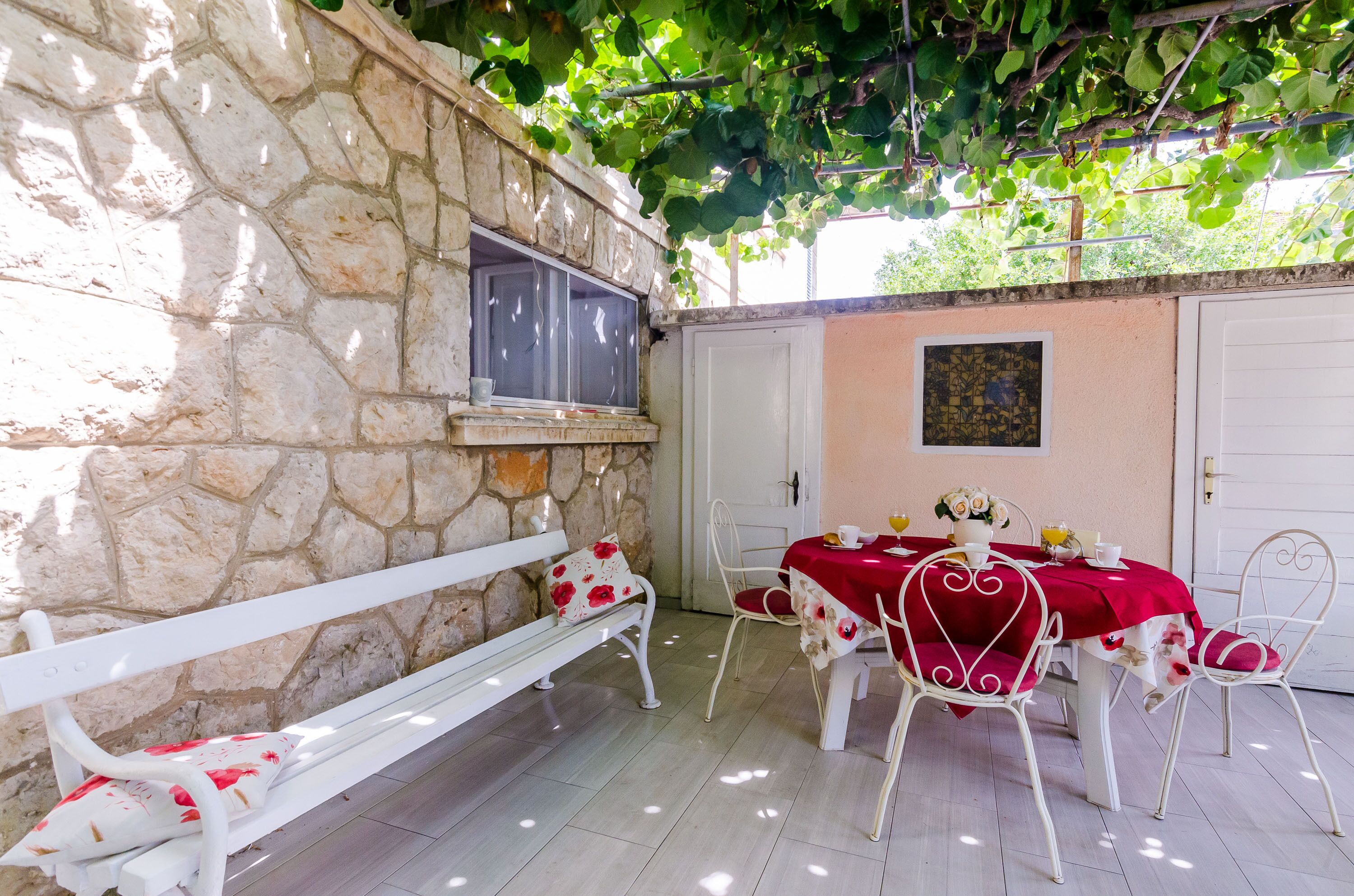 A stone wall with a window, a white bench, a table with a red tablecloth, and white chairs.