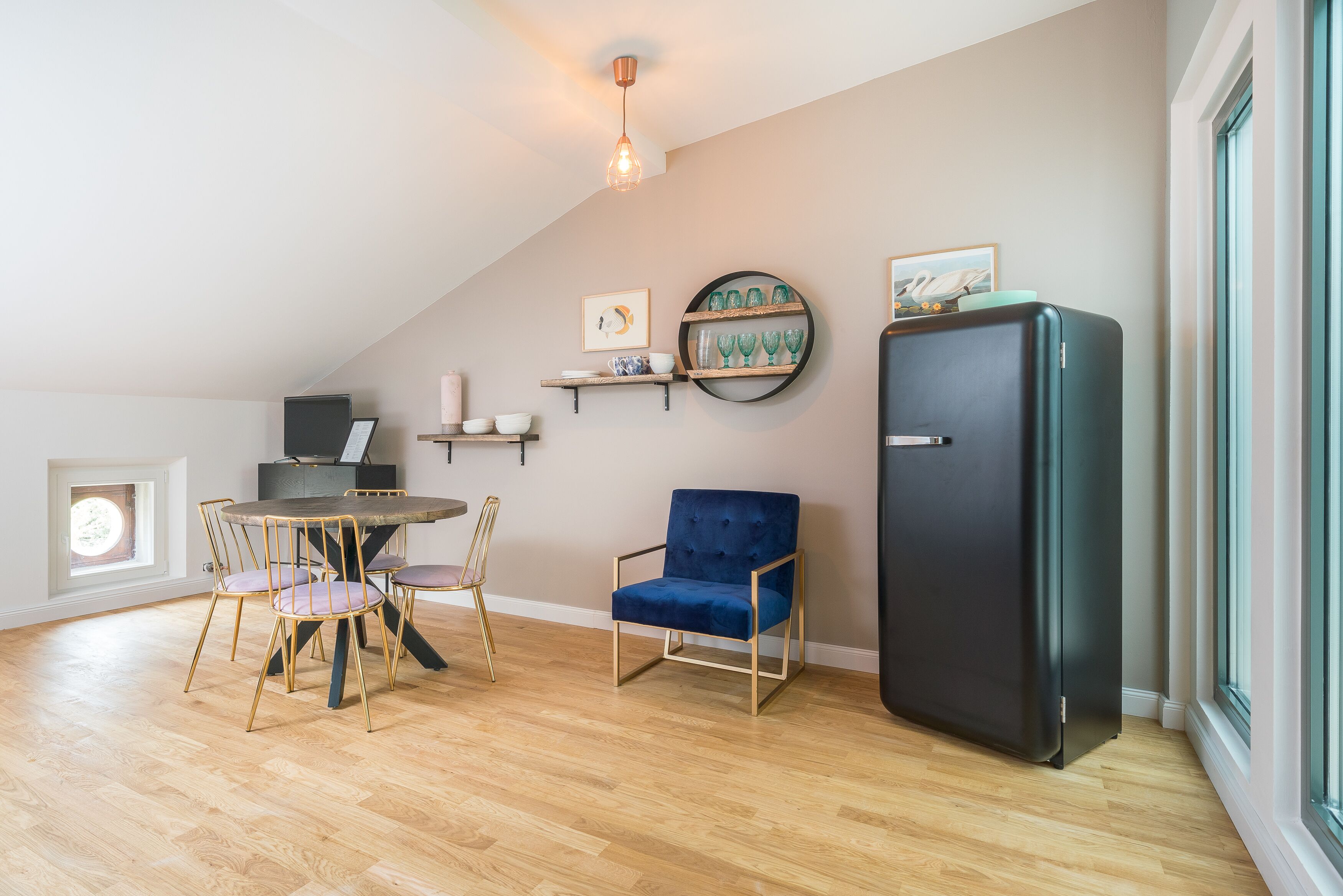 A modern kitchen with a dining table, chairs, a blue armchair, a black refrigerator, and shelves with glassware.