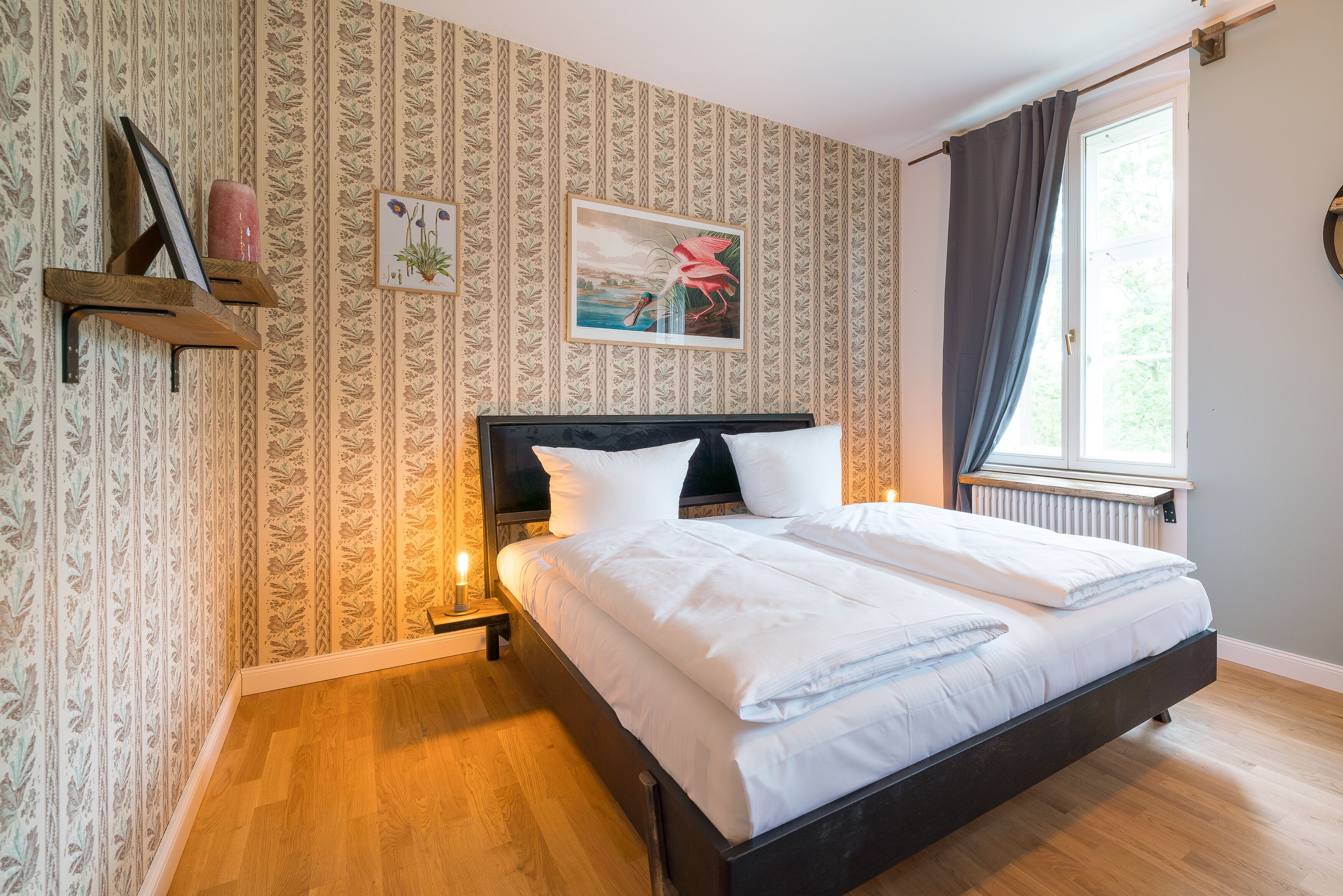 Un lit bien fait, avec du linge de lit blanc, une tête de lit en bois et une table de chevet avec une lampe. La pièce est décorée de papier peint à motifs et possède une fenêtre avec des rideaux.