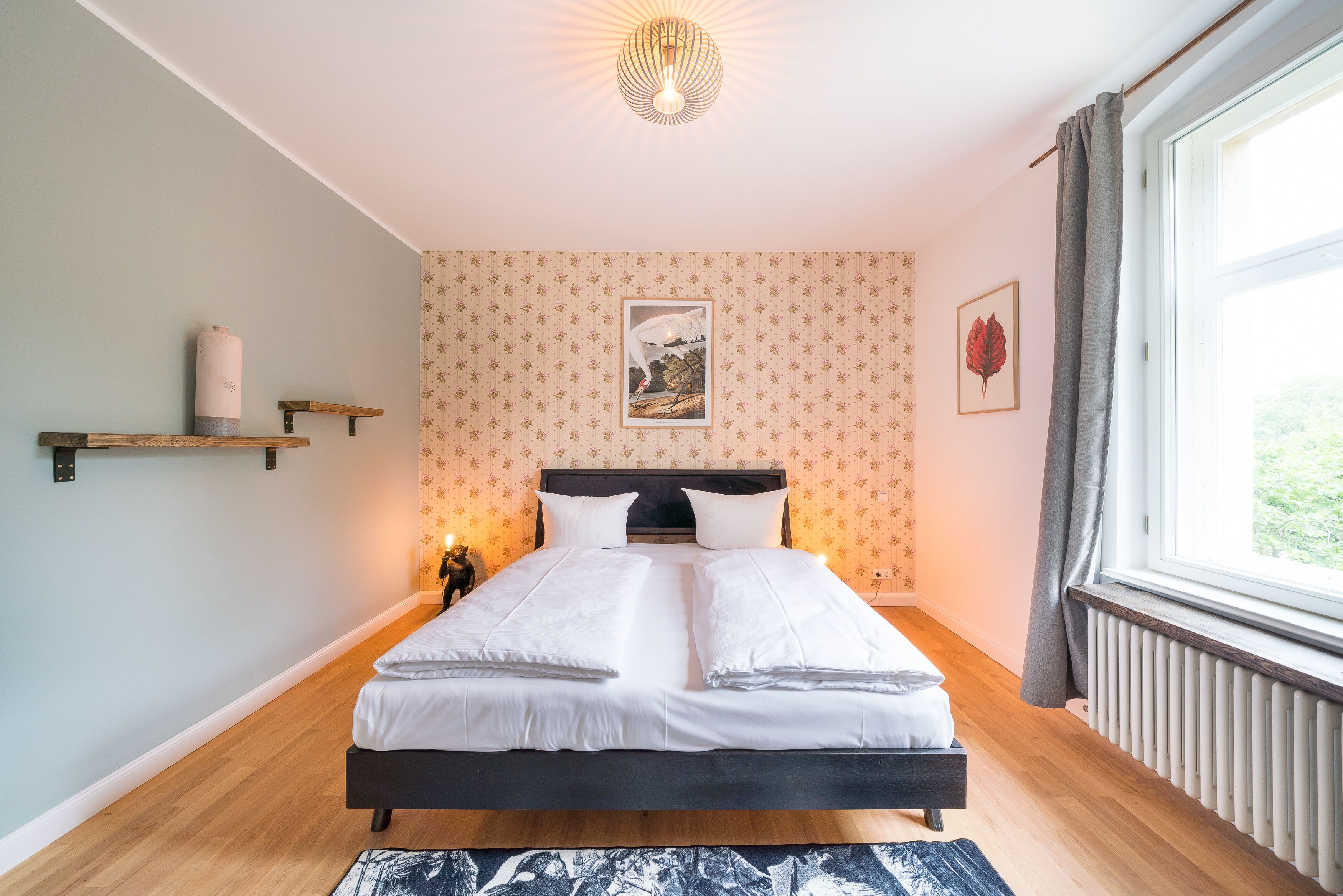 A neatly made bed in a room with a wooden floor, a radiator, and a window with curtains.
