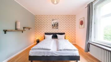 A neatly made bed in a room with a wooden floor, a radiator, and a window with curtains.