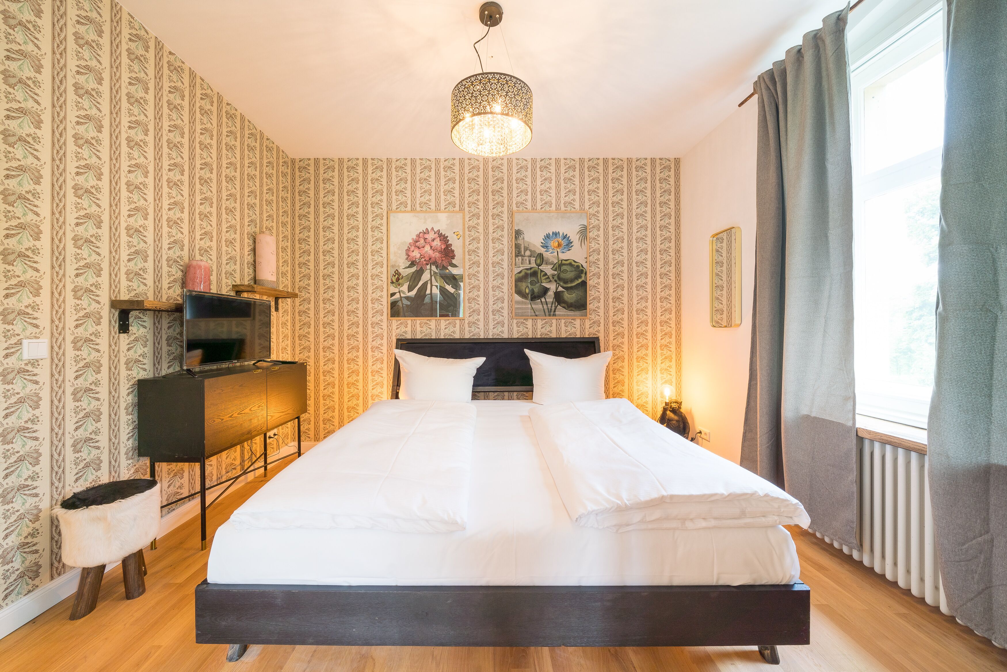 Ein ordentlich gemachtes Bett mit weißen Leinen, einem Holzkopfschild und einem Nachttisch mit Lampenschirm. Der Raum hat raumschichtenes Tapetenmuster und ein großes Fenster mit Vorhängen.