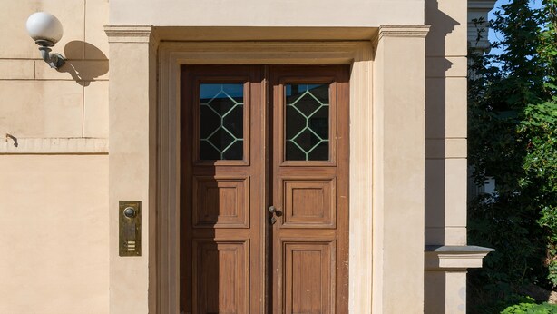 Puerta doble de madera con paneles de vidrio y una placa de latón, en un edificio color beige con base de piedra.