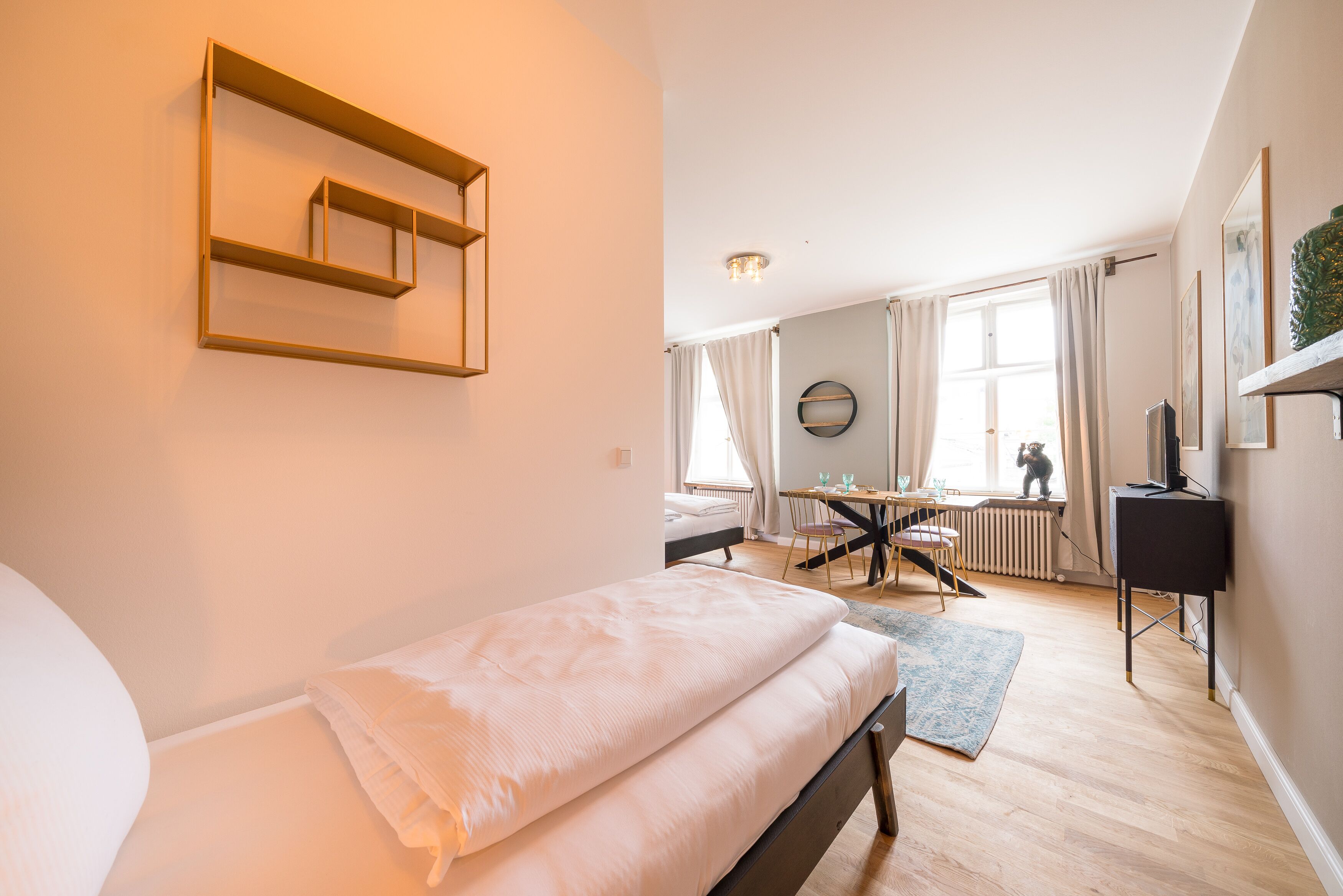 Ein Schlafzimmer mit Bett, einem kleinen Tisch und einem Sessel. Es gibt ein Fenster mit Vorhängen, eine Heizung und einen Fernseher auf einem Schrank.