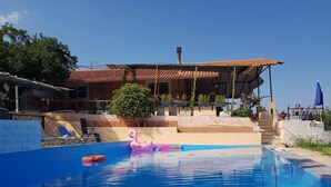 A swimming pool with a pink flamingo float, a covered patio area with outdoor seating, and a view of a building with a tiled roof. - Charming 4-bed House in Pyrgetos (Tempi)